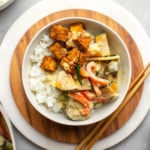 white bowl filled with white rice, crispy tofu, and thai green curry with chopsticks on round wood serving tray