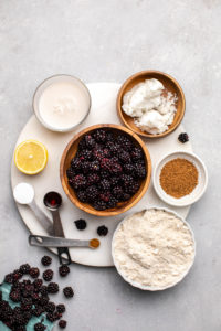 ingredients for blackberry cobbler arranged in small bowls on round white cutting board