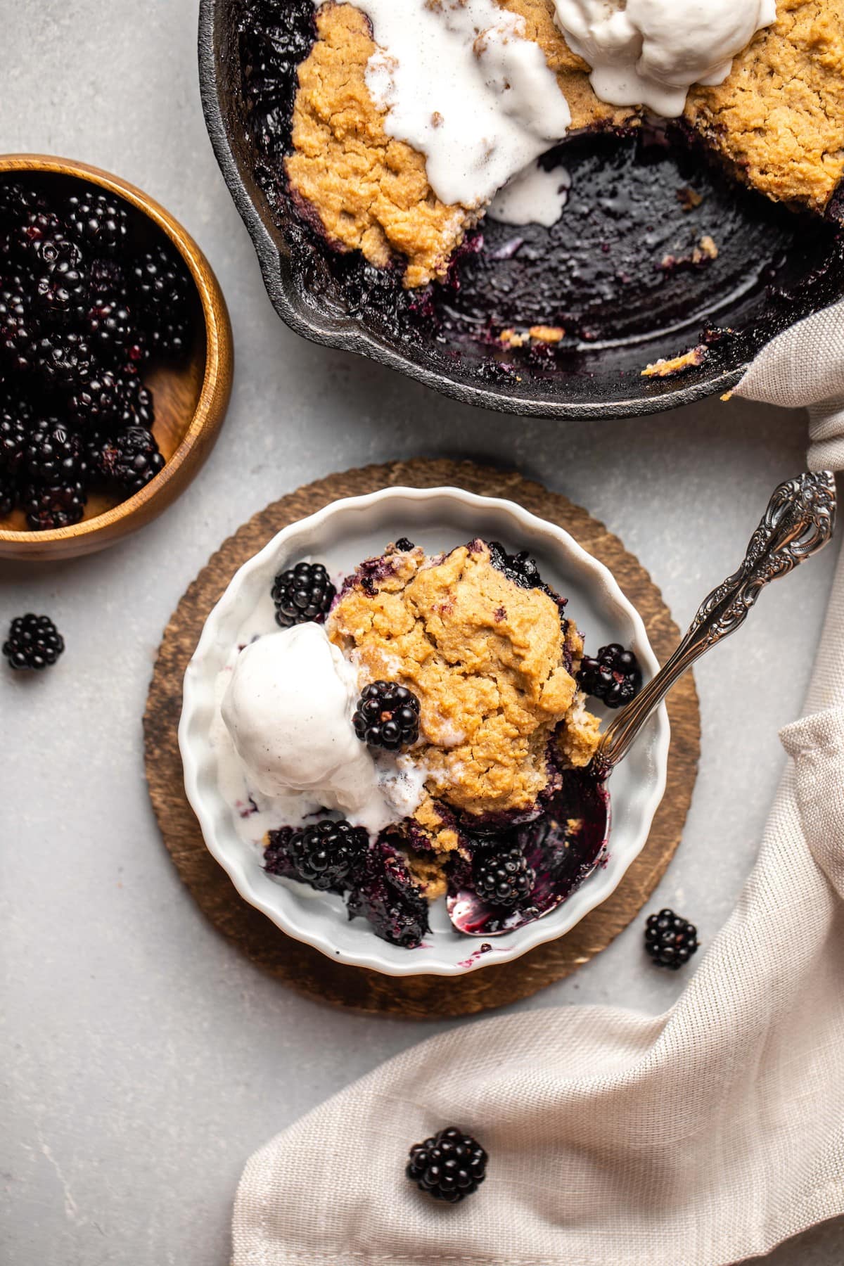 small dish of blackberry cobbler topped with blackberries and vanilla ice cream with skillet of blackberry cobbler in the background