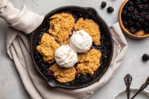 blackberry cobbler in cast iron skillet topped with vanilla ice cream on grey background