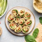 hummus pinwheels on white speckled plate surrounded by tortillas, spinach, and sundried tomatoes