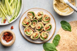 hummus pinwheels on white speckled plate surrounded by tortillas, spinach, and sundried tomatoes