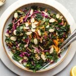everyday kale salad in large white bowl with sercving utensils and lemon wedges