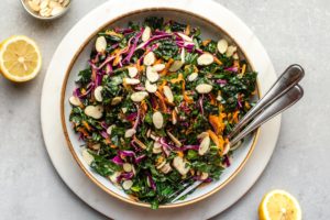 everyday kale salad in large white bowl with sercving utensils and lemon wedges
