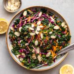 kale salad in large white bowl with lemon wedges and slivered almonds on the side