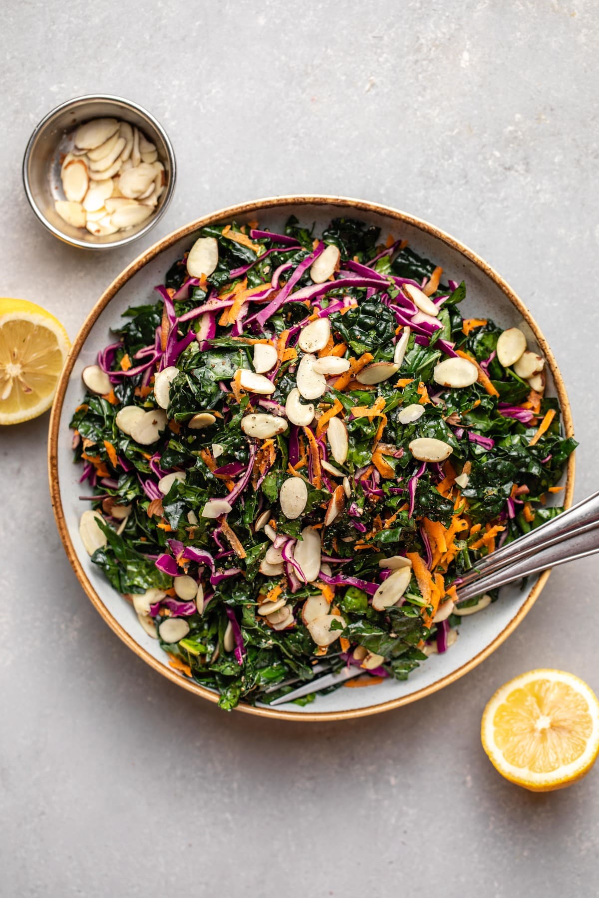 kale salad in large white bowl with lemon wedges and slivered almonds on the side