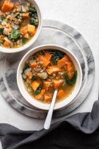 two bowls of sweet potato & kale soup with silver spoons on round marble cutting board