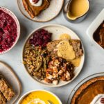 small white plate with lentil loaf, cranberry sauce, and sweet potato casserole surrounded by large thanksgiving dishes on marble background