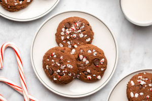 three cookies on a white plate next to candy canes and a glass of milk on white marble background