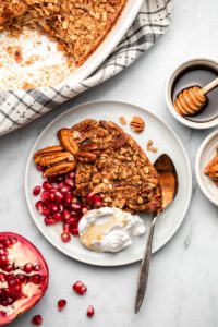 plate of maple pecan baked oatmeal decorated with pecans, pomegranate, seeds, coconut yogurt, and maple syrup on marble background