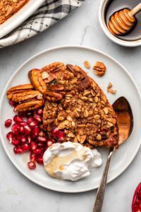 piece of maple pecan baked oatmeal on plate with pecans, pomegranate, coconut yogurt, and maple syrup