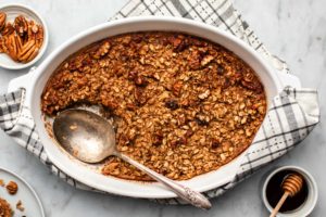 baked maple pecan oatmeal in round white casserole dish with large serving spoon on marble background