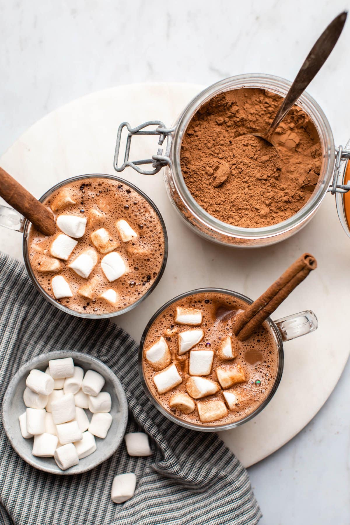 two glass mugs of vegan hot chocolate with glass jar of mix on the side on white marble background