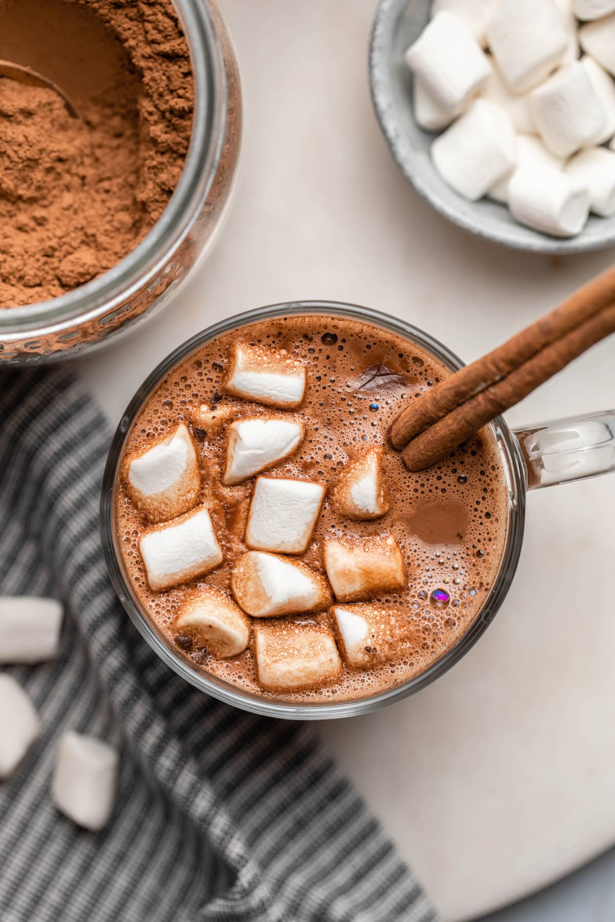 hot chocolate with marshmallows and cinnamon stick in glass mug on white marble background