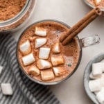 glass mug of hot chocolate with marshmallows and cinnamon stick with glass jar of powdered mix in background