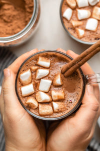 two hands holding glass mug of hot cocoa with marshmallows and cinnamon stick
