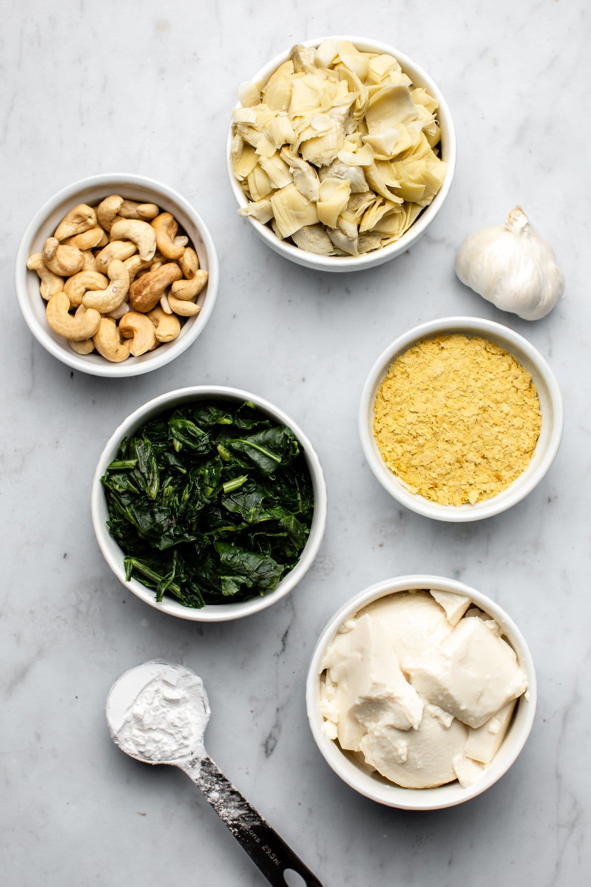 ingredients for spinach and artichoke dip in small white dishes on gray marble background