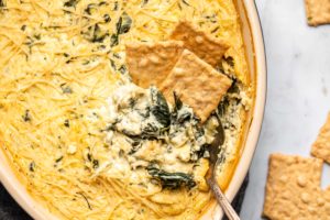three crackers in spinach and artichoke dip with spoon in dish on grey marble background