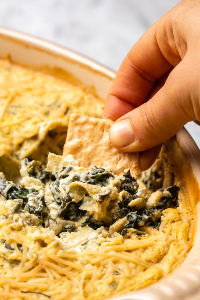 hand dipping cracker into vegan spinach and artichoke dip
