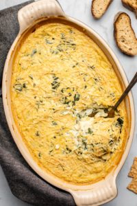 baked spinach and artichoke dip with spoon in it and crostini in background on gray marble background