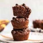 stack of 3 chocolate banana muffins on white plate with bananas in background