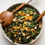 curried kale salad in grey bowl with wood tongs on white background