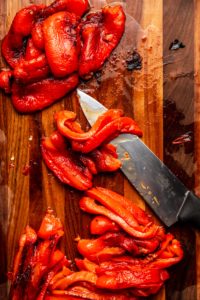 roasted bell peppers being sliced with a knife on wood cutting board