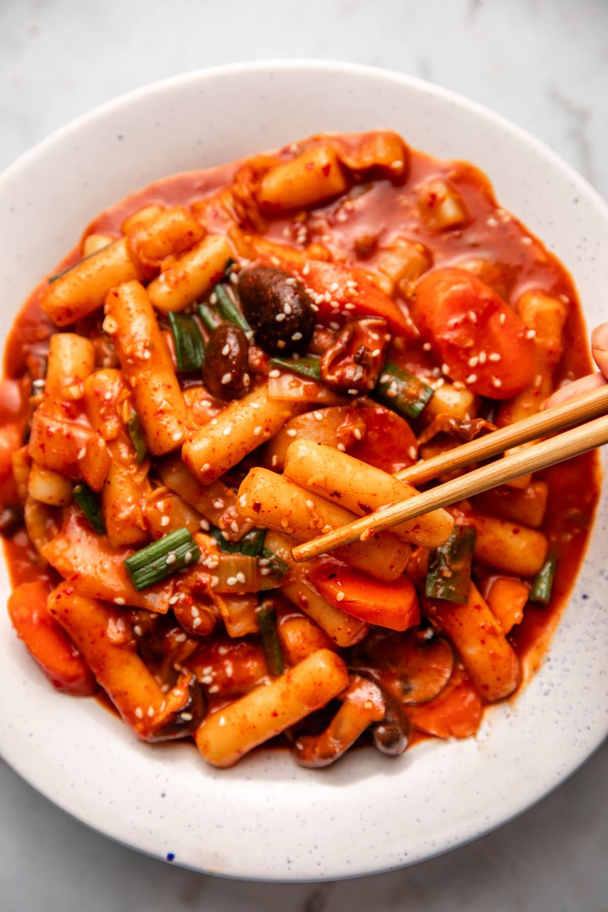 chopsticks holding two saucy rice cakes over bowl of tteokbokki on white marble background