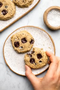 two chocolate chip cookies on white plate with hand grabbing for a cookie on marble background