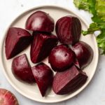 plate of roasted beets next to uncooked beet bulbs and beet greens on marble background