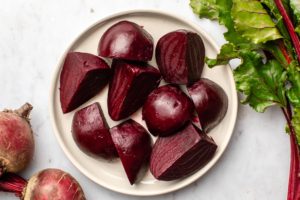 plate of roasted beets next to uncooked beet bulbs and beet greens on marble background