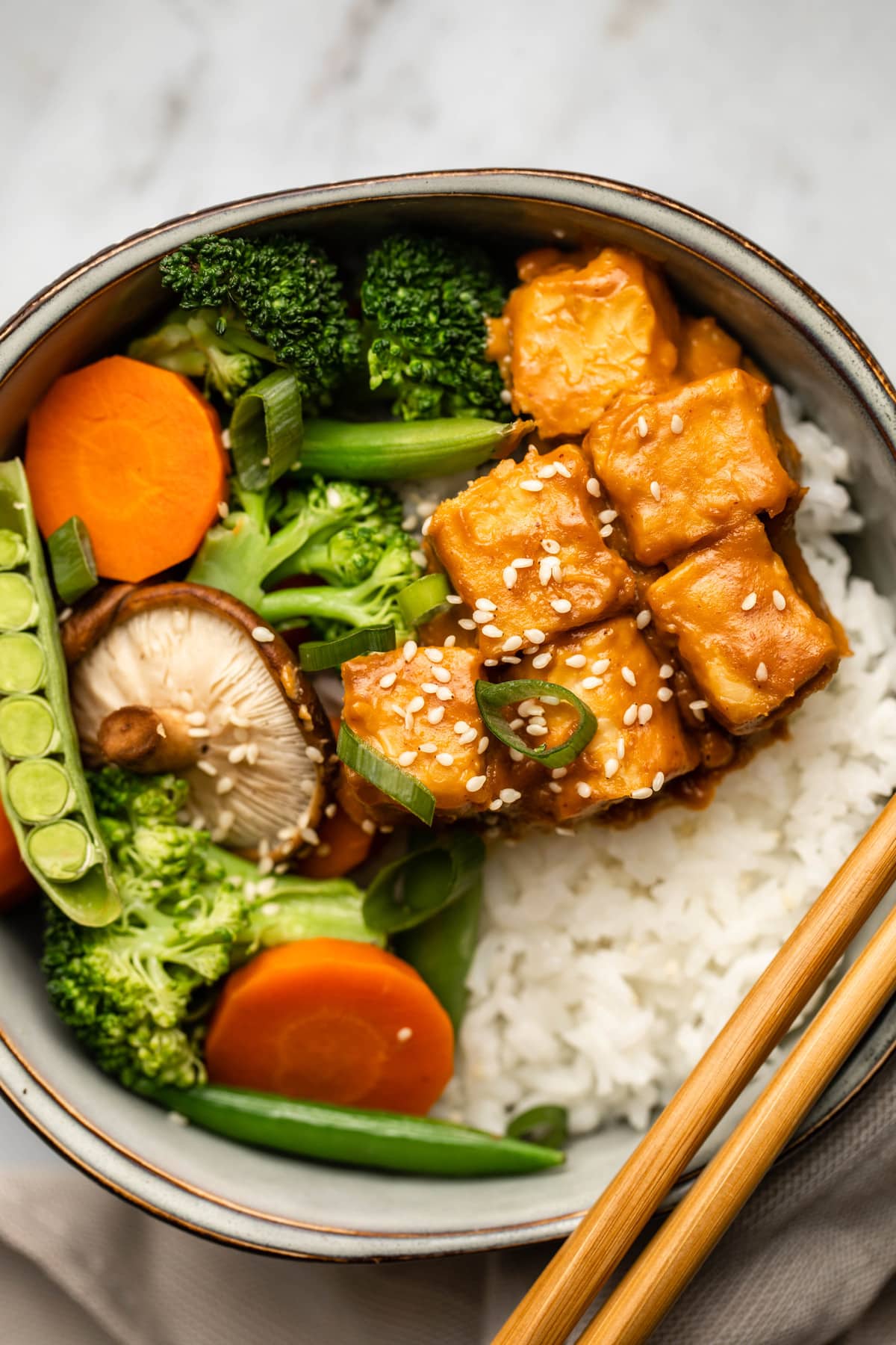 close up image of tempeh, vegetables, and rice in gray bowl with chopsticks