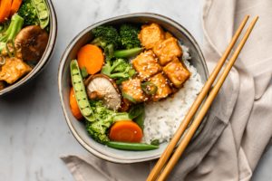bowl of rice, tempeh, and steamed vegetables with chopsticks on marble background