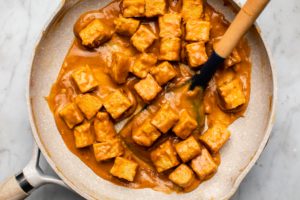 cooked saucy stovetop peanut tempeh in tan saucepan with wooden spatula on marble background
