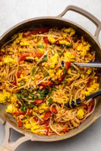 large pan of cooked noodles mixed with scrambled tofu and pad thai sauce on grey background