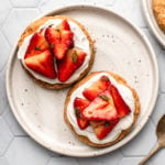 two shortcakes topped with coconut cream and macerated strawberries on white tile background