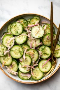 bowl of cucumber dill salad with gold serving utensils on marble background