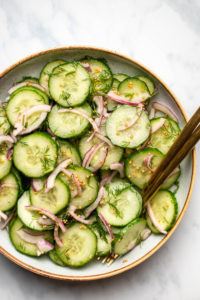 bowl of cucumber dill salad with gold serving utensils on marble background