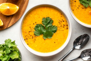 bowl of red lentil soup topped with cilantro and black pepper with side of lemons on grey background