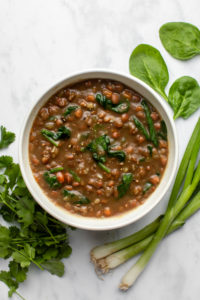 bowl of lentil soup surrounded by cilantro, spinach, and green onions on marble background