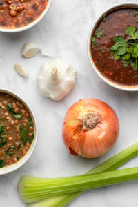 garlic, onion, and celery with bowls of soup on marble background