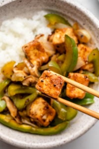 chopsticks holding crispy piece of black pepper tofu over bowl of stir fry and white rice
