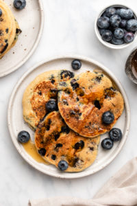 3 blueberry pancakes on white plate topped with fresh blueberries and maple syrup