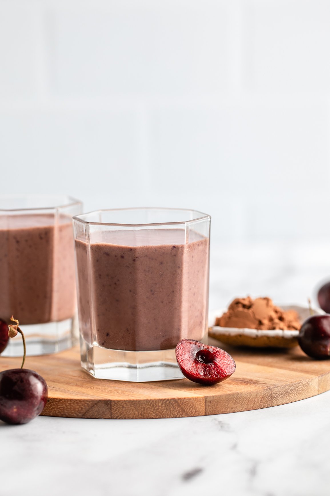 two glasses of chocolate cherry smoothie against white wall with fresh cherries and cacao powder
