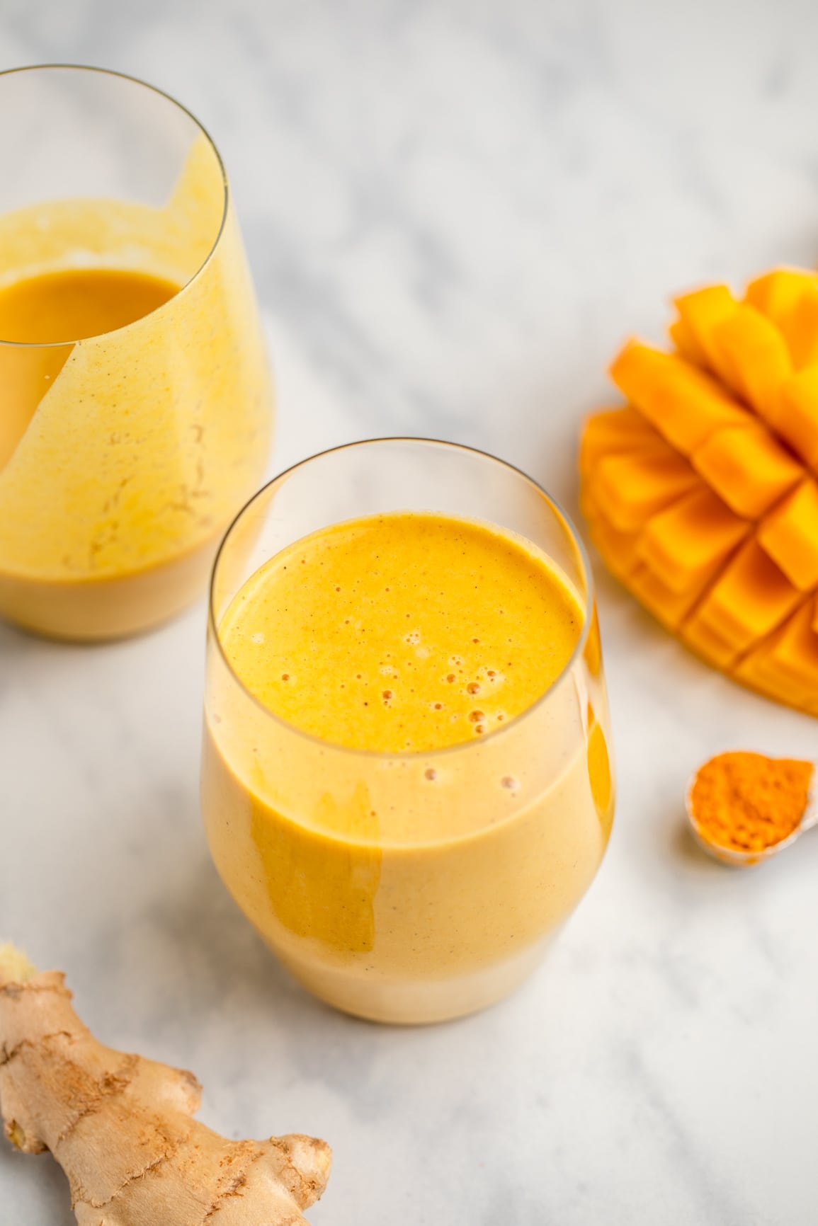 two glasses of golden milk mango smoothie with scored mango on side on marble background