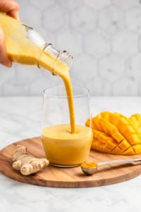 hand pouring golden milk mango smoothie into empty glass on wood serving board