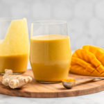 two glasses of golden milk mango smoothie on wood cutting board with sliced mango and fresh ginger on the side