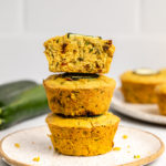 stack of savory zucchini muffins on white plate with zucchini in backgound