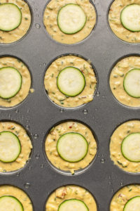 savory zucchini muffin batter in greased muffin tin topped with slice of zucchini