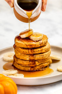 hand pouring maple syrup over stack of pumpkin pancakes topped with banana slices